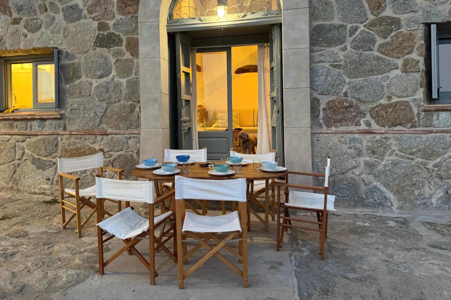 Mesa puesta al atardecer ante la puerta de una casa de piedra, con un perro asomado