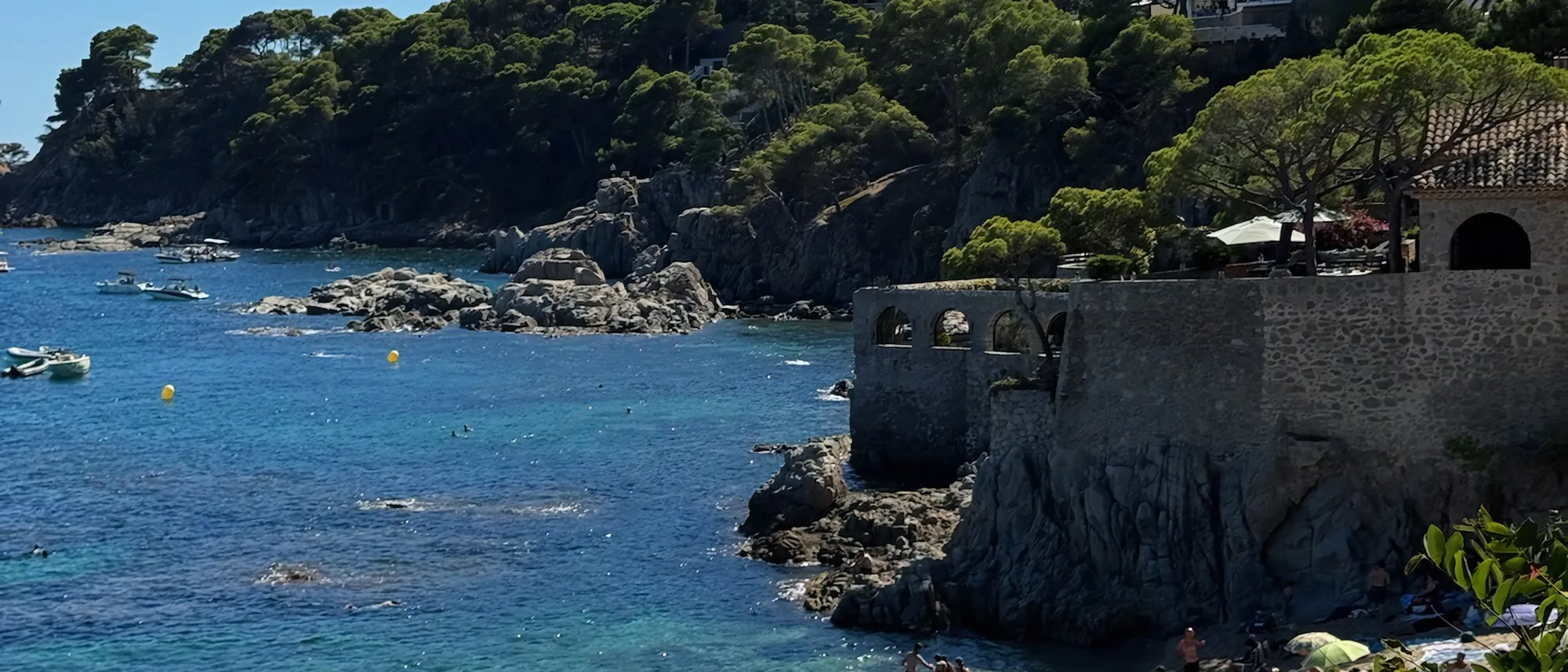 Cala de la Costa Brava con barcas fondeadas, pinos y una terraza de piedra con arcos sobre el agua