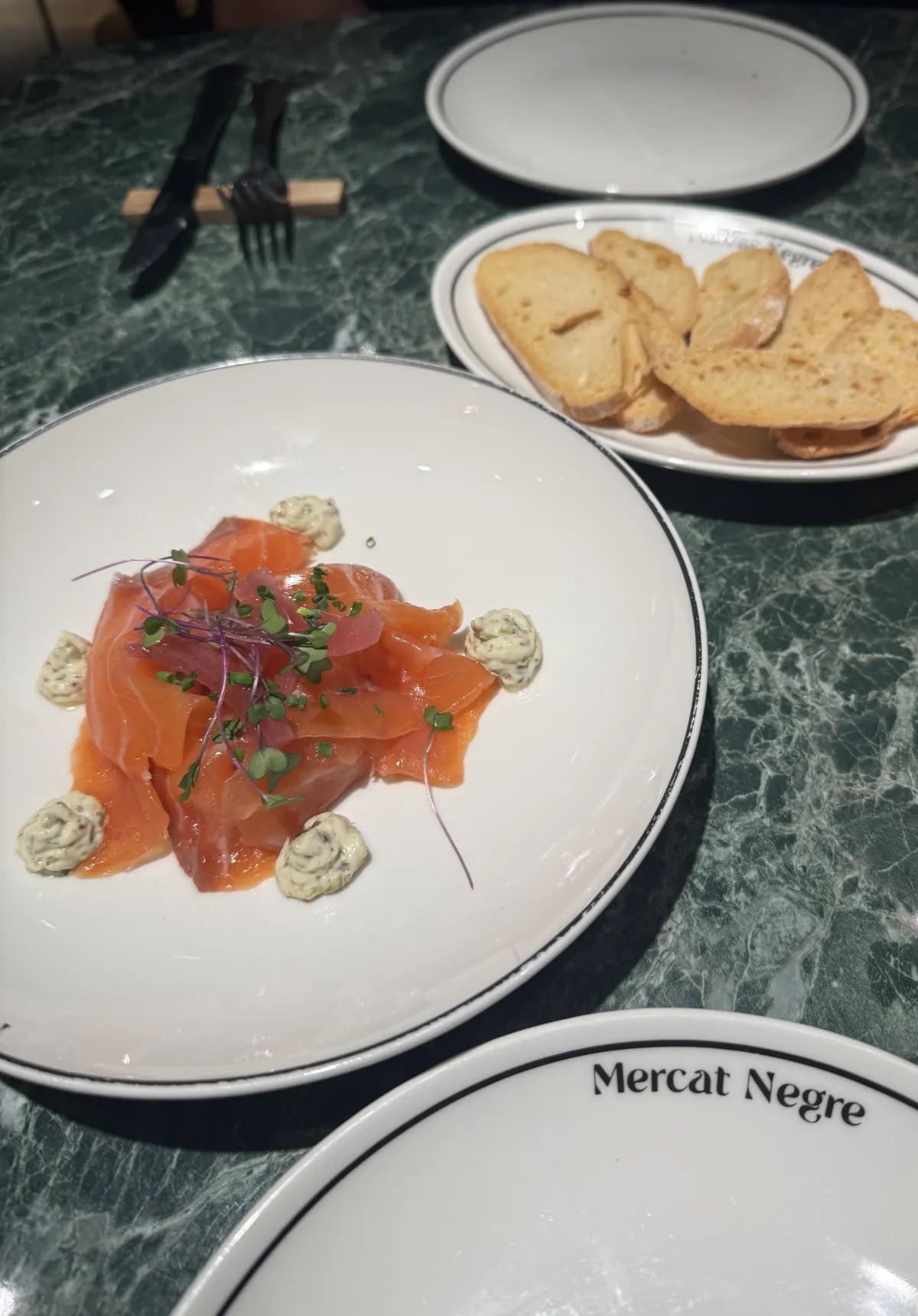 Salmón marinado con brotes tiernos sobre mármol verde, con pan tostado y la vajilla de Mercat Negre