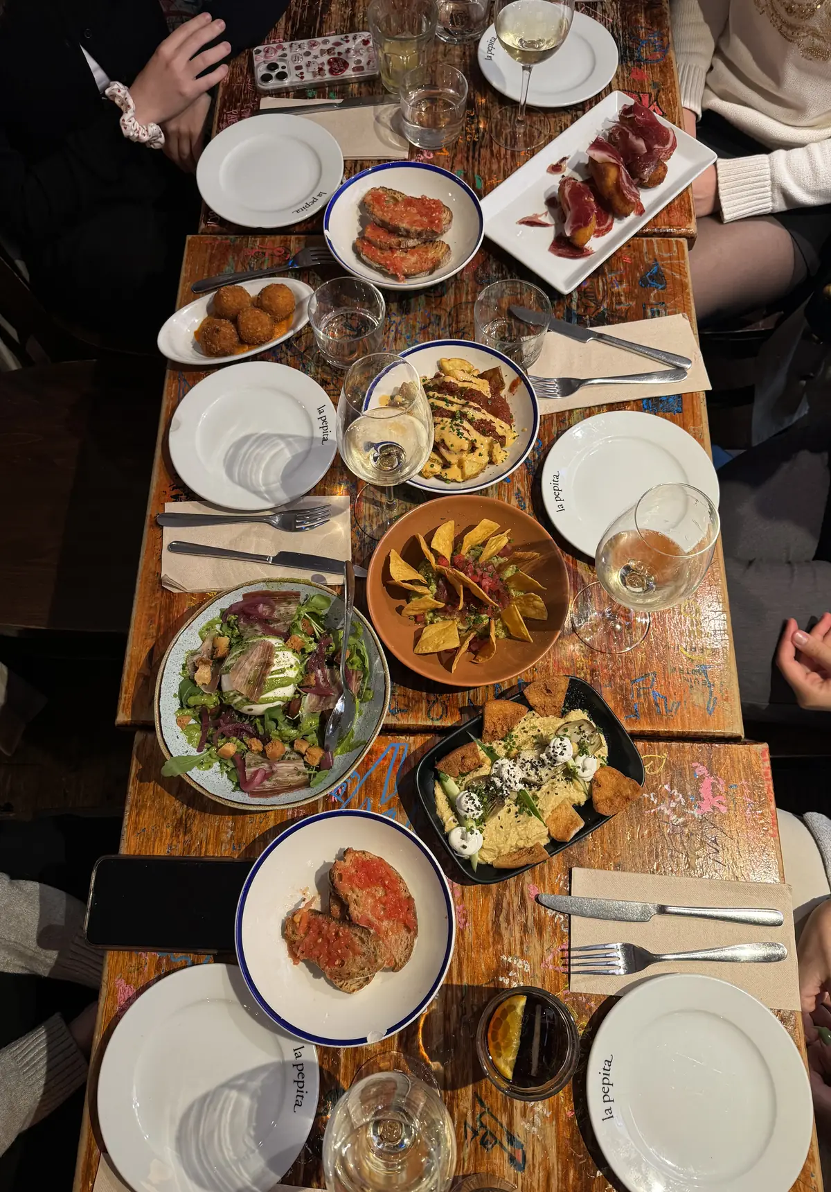 Mesa de madera llena de tapas para compartir, con pan con tomate, croquetas y vino blanco, vista desde arriba